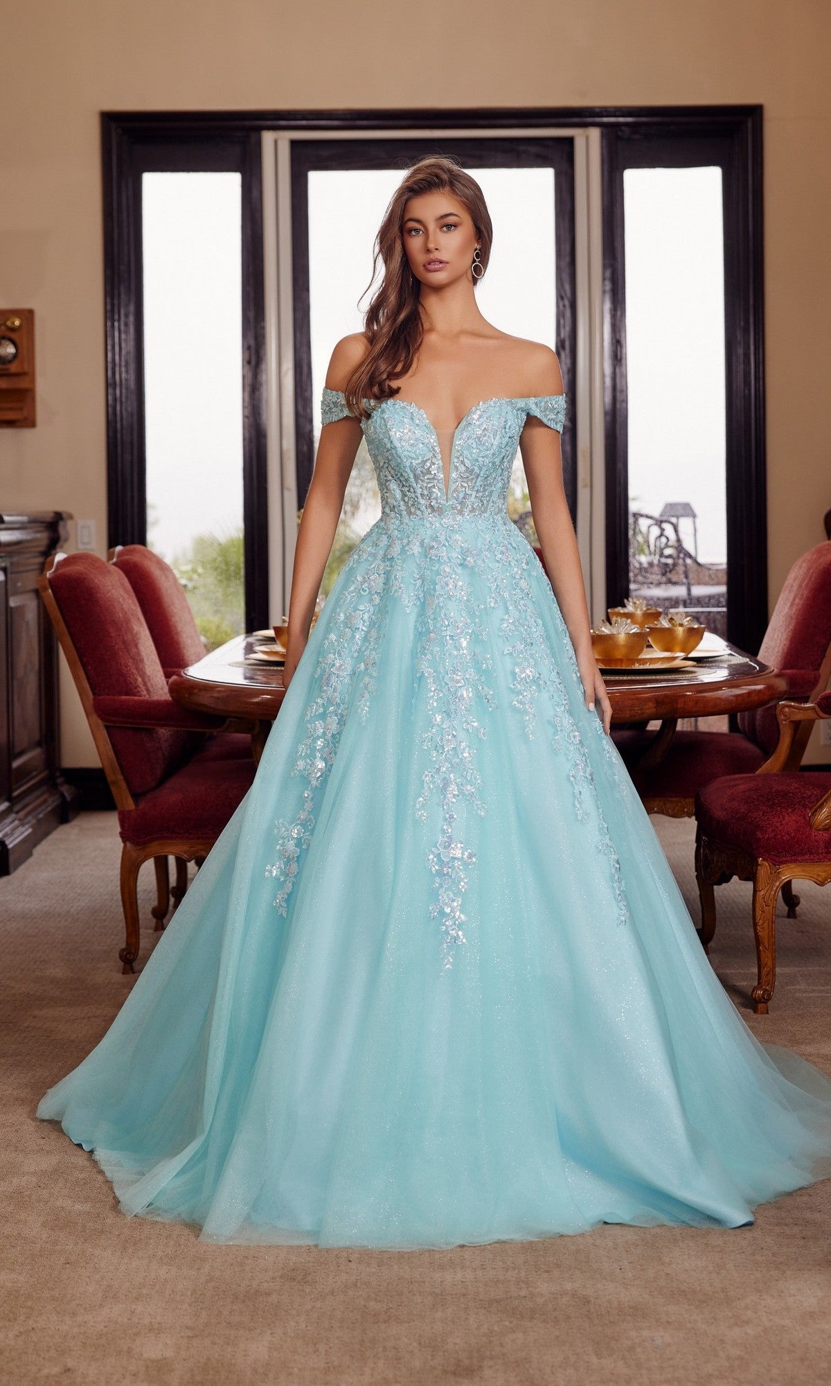 A woman stands indoors by a dining table and large windows, wearing the Nox Anabel H1881—a long prom dress with an off-the-shoulder tulle design and delicate floral embroidery.